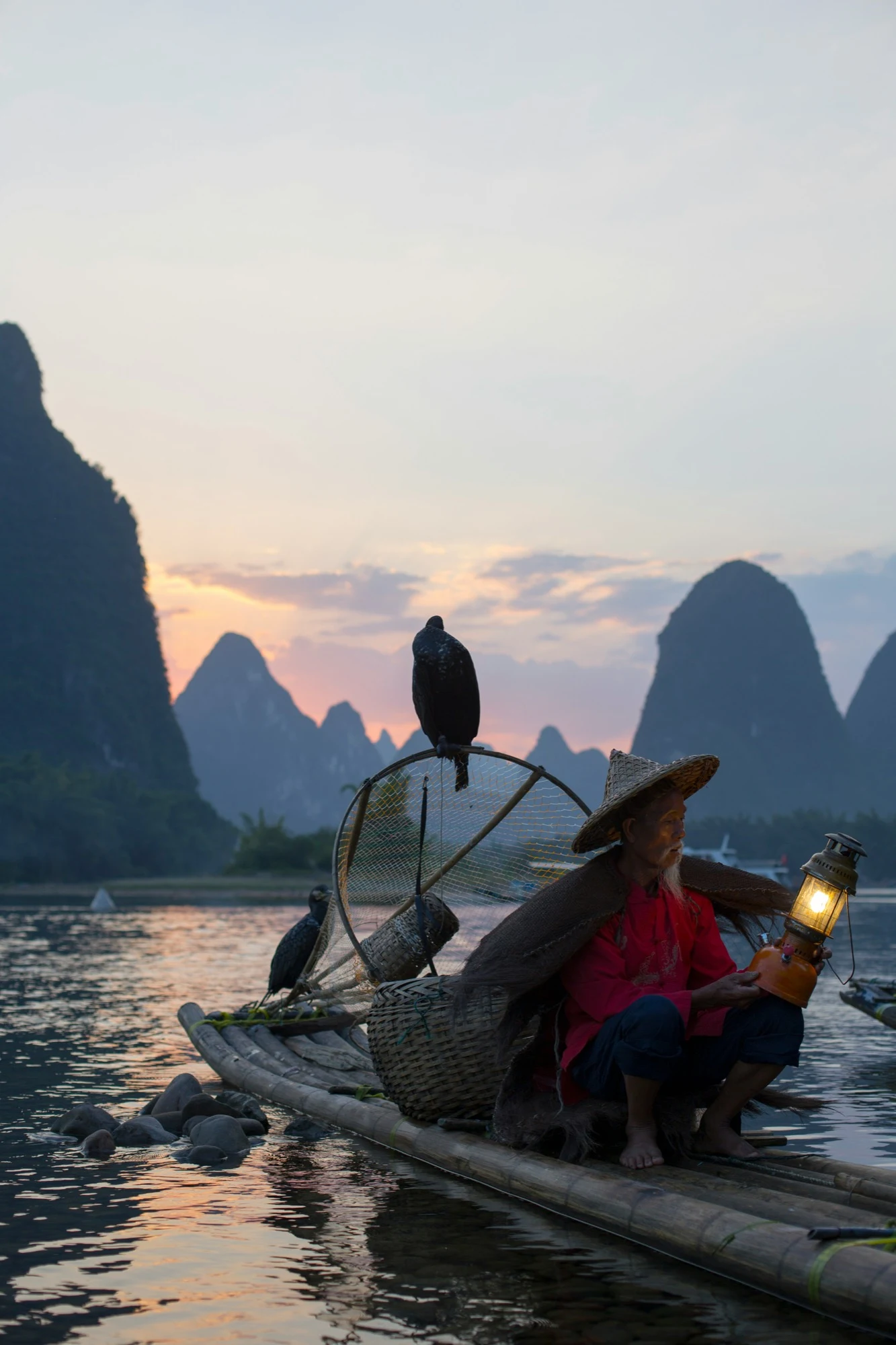 A cormorant fisherman holding a glowing lantern on a bamboo raft on the Li River near Guilin at dusk